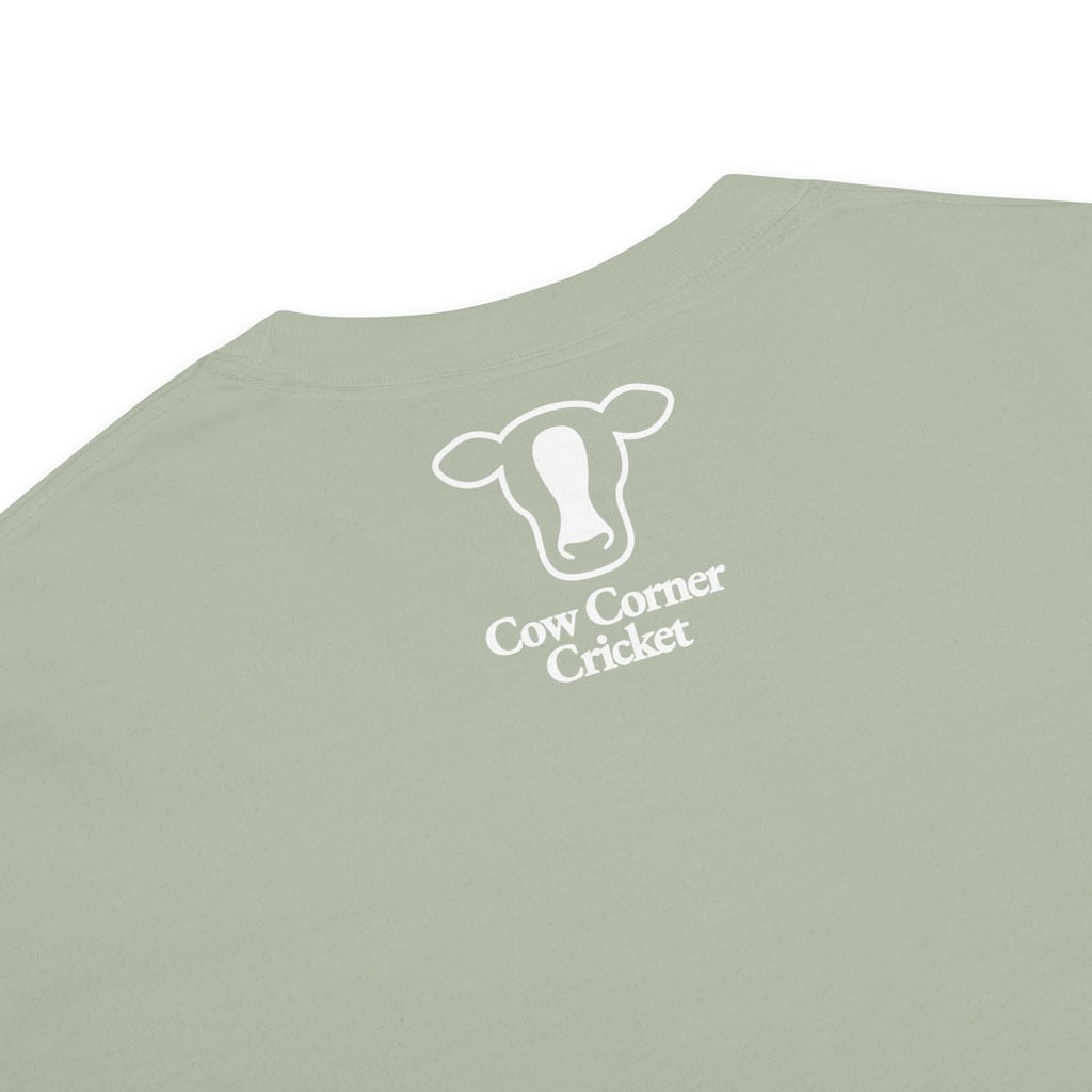 A close-up of a sage green t-shirt  and the text 'Cow Corner Cricket' printed on the center.