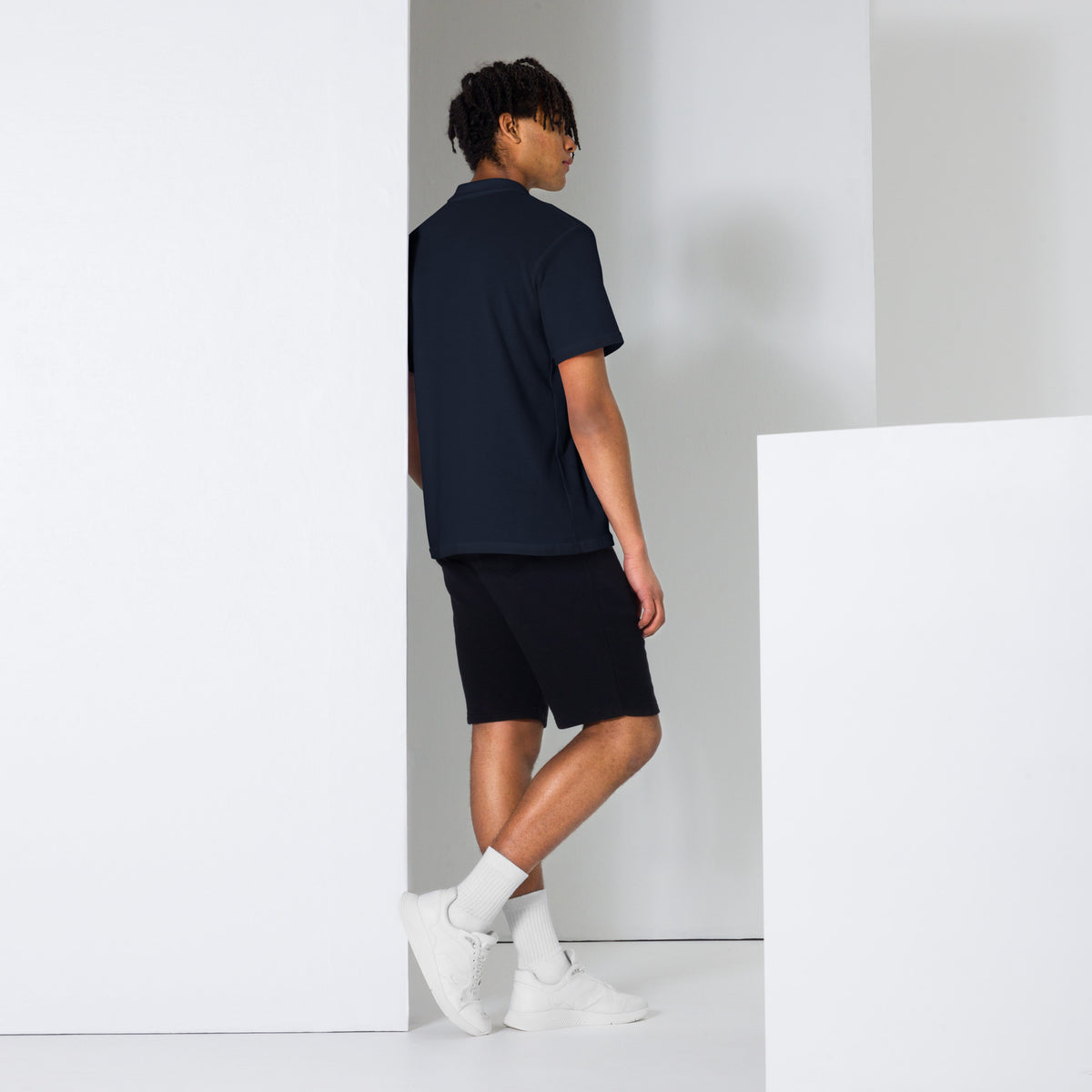 Person wearing a black t-shirt and shorts standing against a white wall.